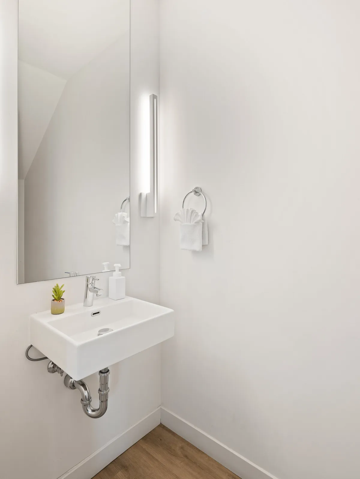 A serene bathroom space featuring a sink, mirror, modern light fixture, a small plant, and a towel neatly arranged. The uncluttered design creates an inviting nook for self-care and refreshment.