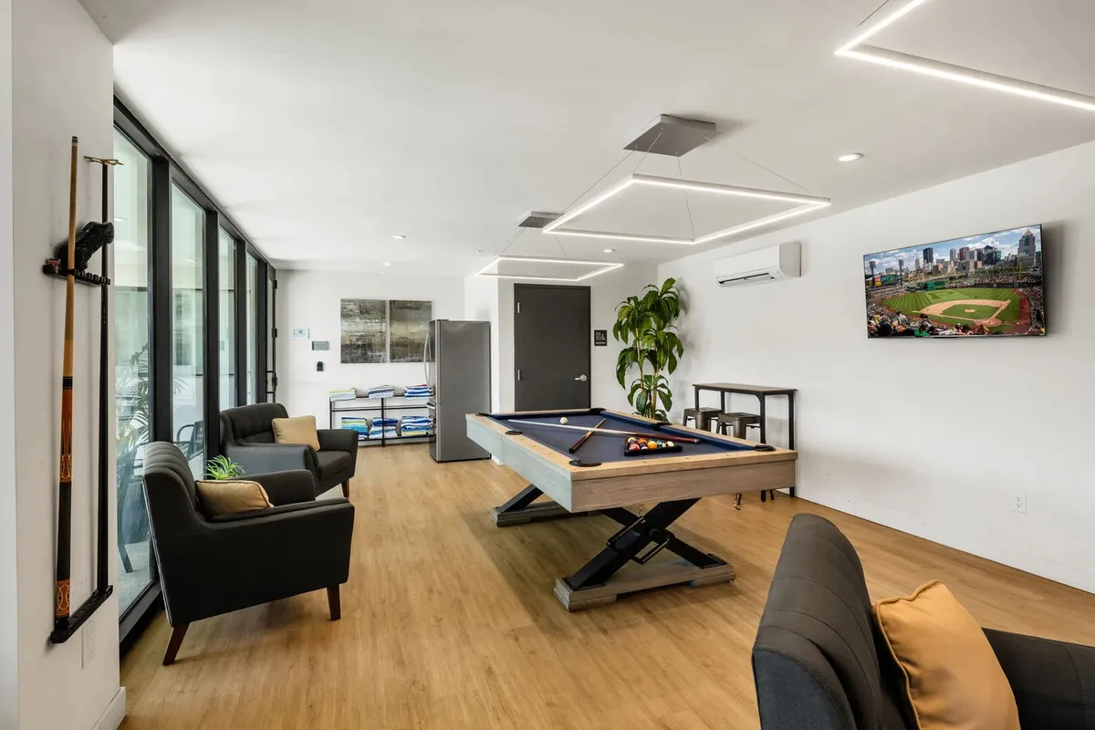 An inviting view of a stylish community clubhouse interior, highlighted by a pool table, plush sofas, a TV, and greenery. The space exudes a relaxed, social atmosphere perfect for unwinding or friendly competition after a night out.