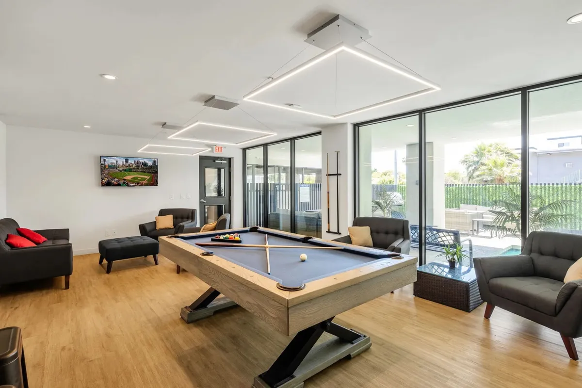A spacious Community Room featuring a modern pool table as the centerpiece, surrounded by comfortable seating, large windows letting in abundant natural light, and a television. The room is set up for both friendly competition and relaxed socializing, with guests enjoying games, conversations, and entertainment.