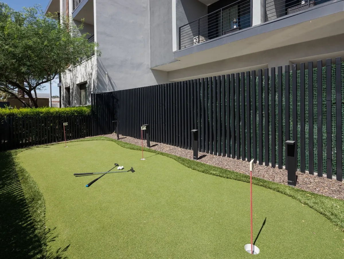 A playful exterior putting green set against a backdrop of modern architecture and lush greenery. Golf clubs can be seen ready for use, and the area is designed for both casual golf practice and friendly competitions among guests. The space encourages outdoor activity and social interaction.