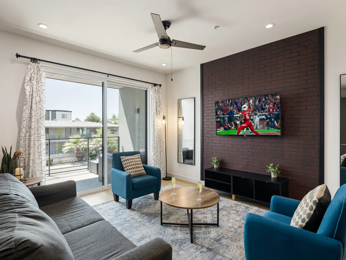 This inviting living room features a sleek modern sectional sofa and stylish blue accent chairs arranged around a round coffee table, with a large TV displaying a sports game, creating a vibrant and comfortable setting for gathering with friends or family.