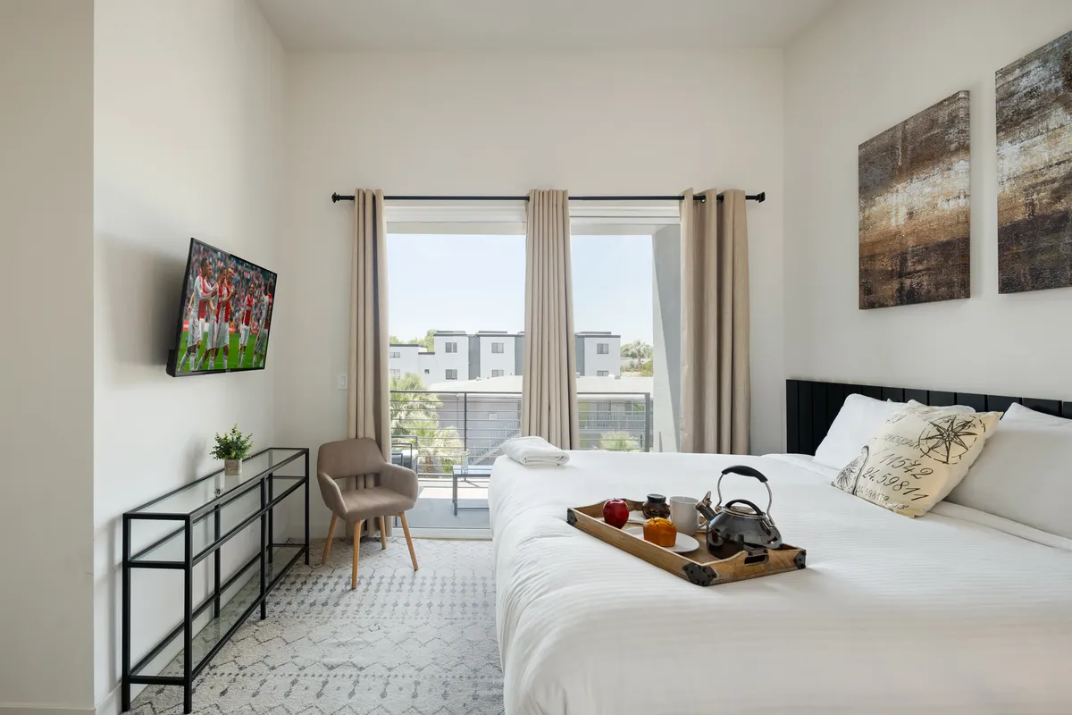 A comfortable bedroom scene with a king bed dressed in soft linens, a tray with breakfast items, a large window letting in natural light, and a TV displaying sports content. The setup feels both inviting and ideal for leisurely mornings or cozy evenings.