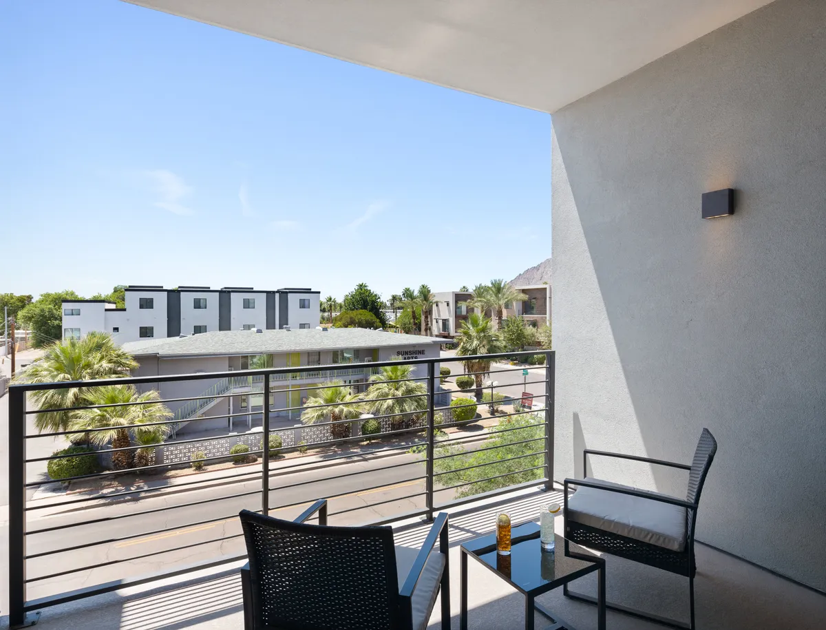 A private balcony setup with comfortable seating overlooks palm trees and distant mountain views. The scene is bathed in soft, natural light, exuding tranquility and inviting guests to enjoy peaceful moments outdoors. The balcony feels intimate, perfect for morning coffee or evening relaxation.